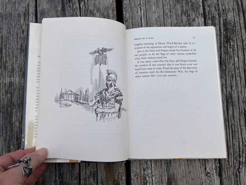 May include: An open book with white pages featuring black ink text and a line drawing of a Roman soldier holding a flag pole with an eagle topper. The text on the right page is titled Origin of a Flag and discusses national symbols.