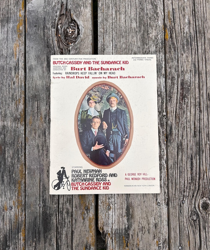 K&ouml;nnte beinhalten: Ein Notenheft f&uuml;r den Film Butch Cassidy und Sundance Kid, komponiert von Burt Bacharach. Das Cover zeigt ein zentrales ovales Portr&auml;t von Paul Newman, Robert Redford und Katharine Ross, erg&auml;nzt durch roten und schwarzen Text.