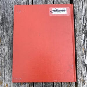 May include: A rectangular hardcover book with a solid orange-red textured cover. In the upper right corner, a white rectangular library sticker is affixed, featuring a black barcode and the printed text Wallace Elementary Library T 21773.