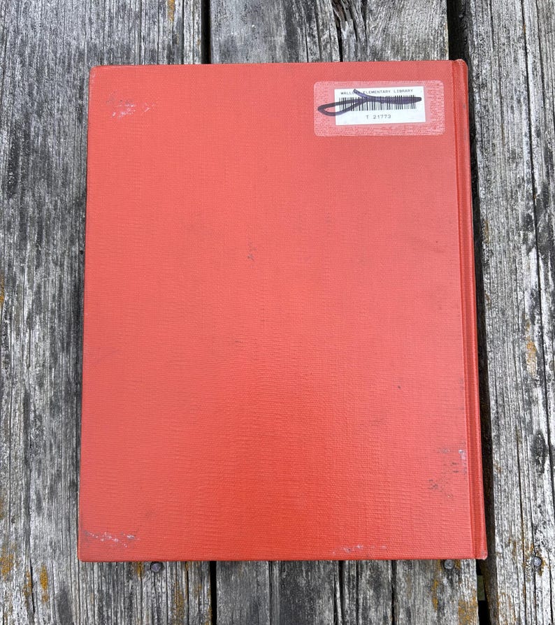 May include: A rectangular hardcover book with a solid orange-red textured cover. In the upper right corner, a white rectangular library sticker is affixed, featuring a black barcode and the printed text Wallace Elementary Library T 21773.
