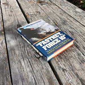May include: A hardcover book titled "FASTNET, FORCE 10" by John Rousmaniere, resting on a weathered wooden surface. The book's cover features a blue background with white and yellow text, and an image of a sailboat in a storm.