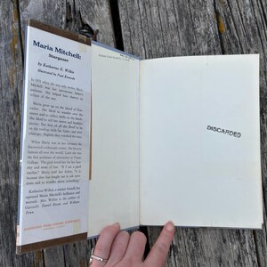 Puede incluir: Un libro de tapa dura titulado Maria Mitchell: Stargazer de Katherine E. Wilkie, que presenta una solapa de sobrecubierta con texto biogr&aacute;fico y una guarda de color blanco roto a la derecha con la palabra DISCARDED estampada en tinta negra.