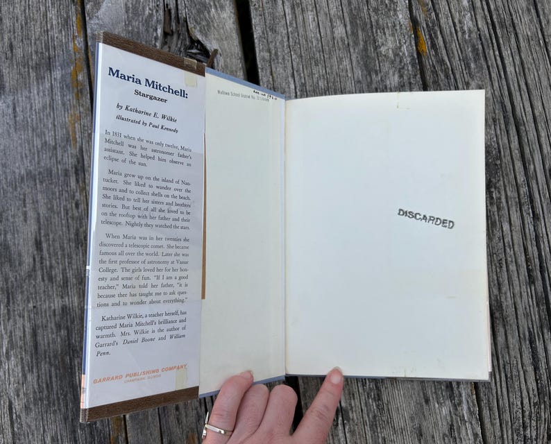 Puede incluir: Un libro de tapa dura titulado Maria Mitchell: Stargazer de Katherine E. Wilkie, que presenta una solapa de sobrecubierta con texto biogr&aacute;fico y una guarda de color blanco roto a la derecha con la palabra DISCARDED estampada en tinta negra.
