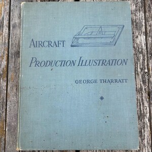 Pu&ograve; includere: Un libro con copertina rigida intitolato Aircraft Production Illustration di George Tharratt con una copertina grigio-azzurra chiara. La copertina presenta un disegno al tratto blu scuro di uno strumento da disegno e il titolo in caratteri serif.