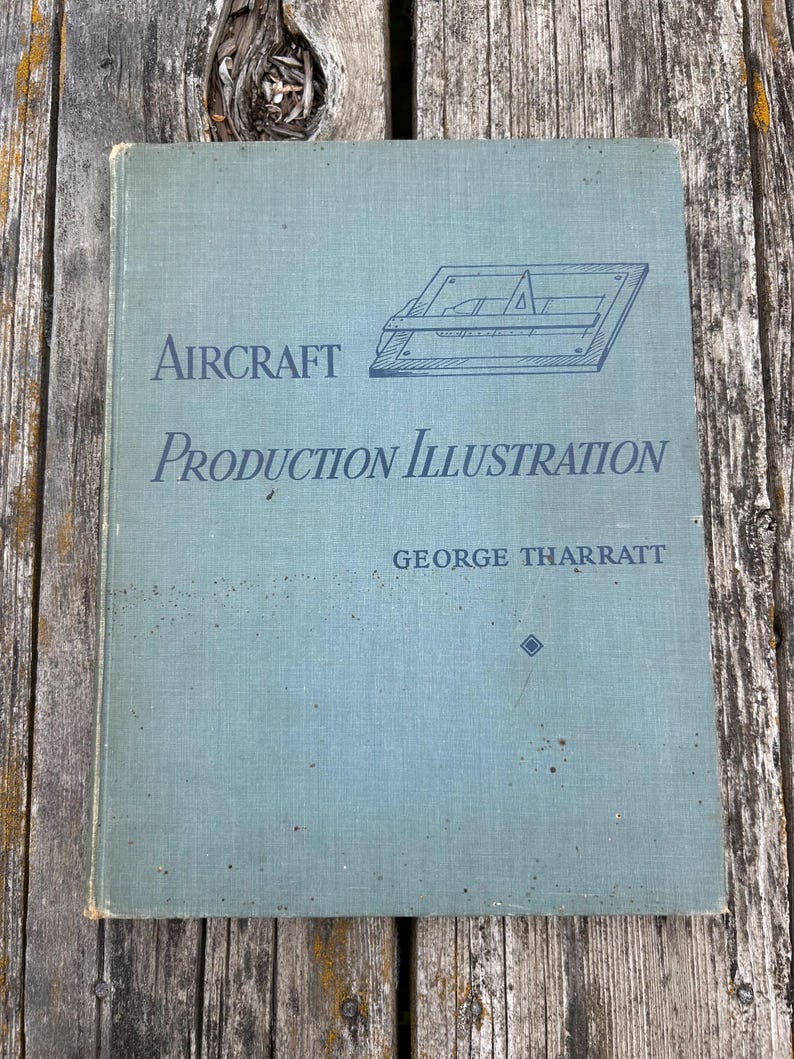 Pu&ograve; includere: Un libro con copertina rigida intitolato Aircraft Production Illustration di George Tharratt con una copertina grigio-azzurra chiara. La copertina presenta un disegno al tratto blu scuro di uno strumento da disegno e il titolo in caratteri serif.