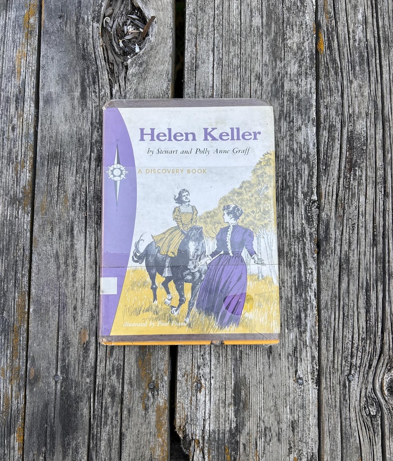 May include: A hardback book titled Helen Keller by Stewart and Polly Anne Graff, A Discovery Book. The cover features a vintage illustration of a person riding a black horse and another person standing in a field, with purple and yellow accents.