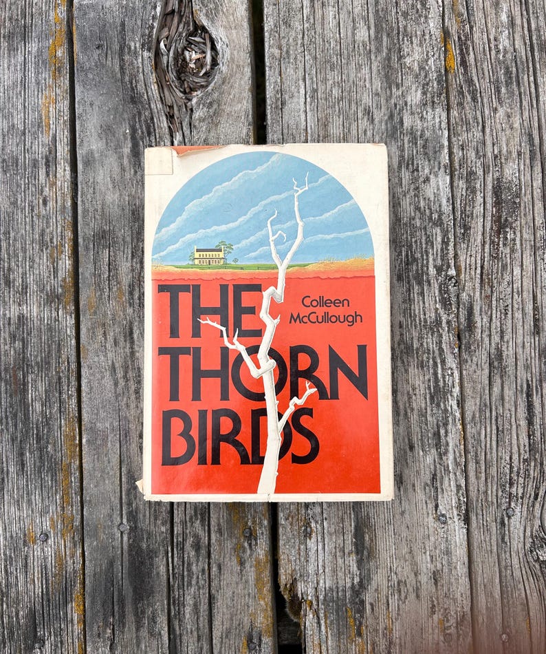 May include: A paperback book titled The Thorn Birds by Colleen McCullough. The cover features a white, leafless tree against a blue sky and a red landscape with a small house. The title is printed in large black letters over the red background.
