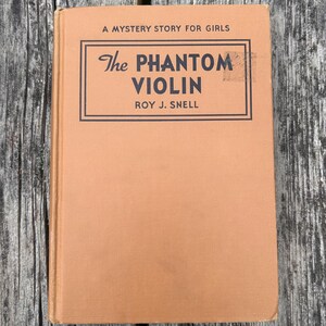 K&ouml;nnte beinhalten: Ein gebundenes Buch mit einem beigefarbenen Stoffeinband mit dem Titel The Phantom Violin von Roy J. Snell, A Mystery Story for Girls. Das Cover zeigt schwarzen Text in einem rechteckigen Rahmen, mit sichtbarem Buchr&uuml;cken und Textur.