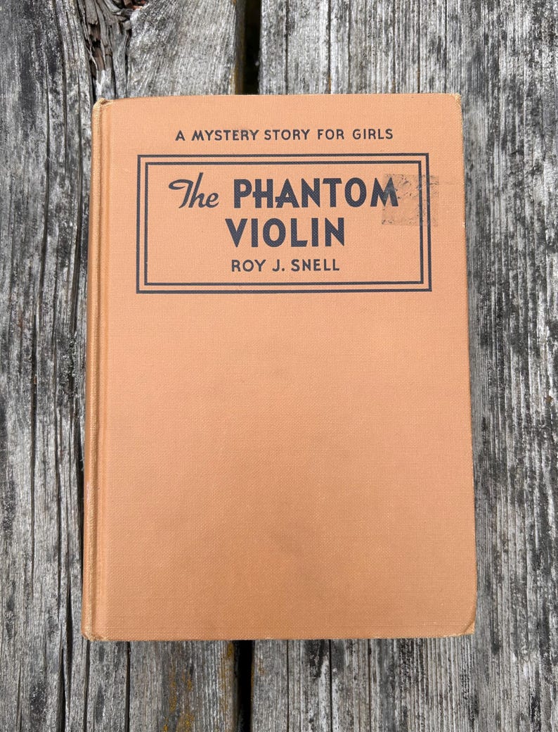 K&ouml;nnte beinhalten: Ein gebundenes Buch mit einem beigefarbenen Stoffeinband mit dem Titel The Phantom Violin von Roy J. Snell, A Mystery Story for Girls. Das Cover zeigt schwarzen Text in einem rechteckigen Rahmen, mit sichtbarem Buchr&uuml;cken und Textur.