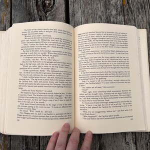 May include: An open paperback book with cream-colored pages featuring black printed text. The left page shows page number 598 and the right page shows page number 599. The book is held open, displaying two columns of narrative text in English.