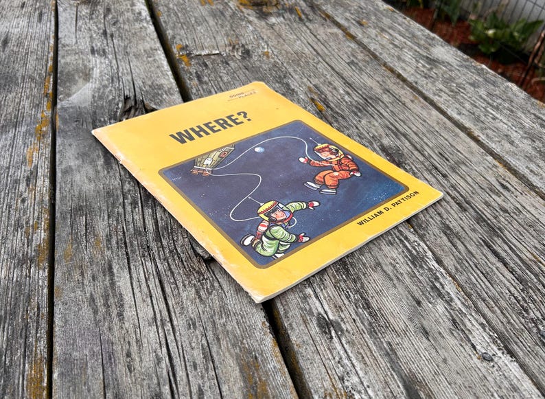 May include: A yellow softcover book titled WHERE? by William D. Pattison. The cover features a dark blue rectangular illustration of two astronauts in orange and green suits floating in space, connected by a tether to a small spacecraft.