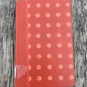 May include: A rectangular hardcover book with a textured red cover featuring a repeating pattern of light pink sunburst icons. A vertical decorative band runs along the left side, and a small white rectangular label is affixed to the bottom left corner.