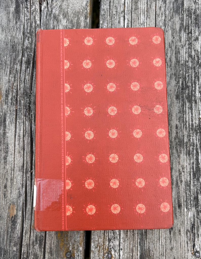 May include: A rectangular hardcover book with a textured red cover featuring a repeating pattern of light pink sunburst icons. A vertical decorative band runs along the left side, and a small white rectangular label is affixed to the bottom left corner.