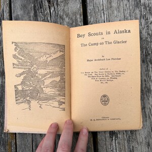 K&ouml;nnte beinhalten: Ein aufgeschlagenes altes Buch mit dem Titel Boy Scouts in Alaska or The Camp on The Glacier von Major Archibald Lee Fletcher. Die linke Seite zeigt eine schwarz-wei&szlig;e Illustration einer Person in einer Schneelandschaft, die rechte Seite Text.