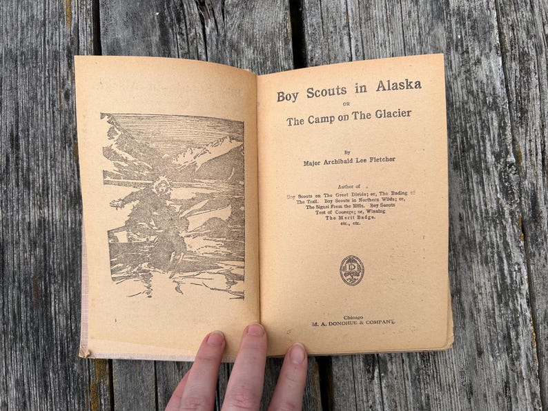 K&ouml;nnte beinhalten: Ein aufgeschlagenes altes Buch mit dem Titel Boy Scouts in Alaska or The Camp on The Glacier von Major Archibald Lee Fletcher. Die linke Seite zeigt eine schwarz-wei&szlig;e Illustration einer Person in einer Schneelandschaft, die rechte Seite Text.