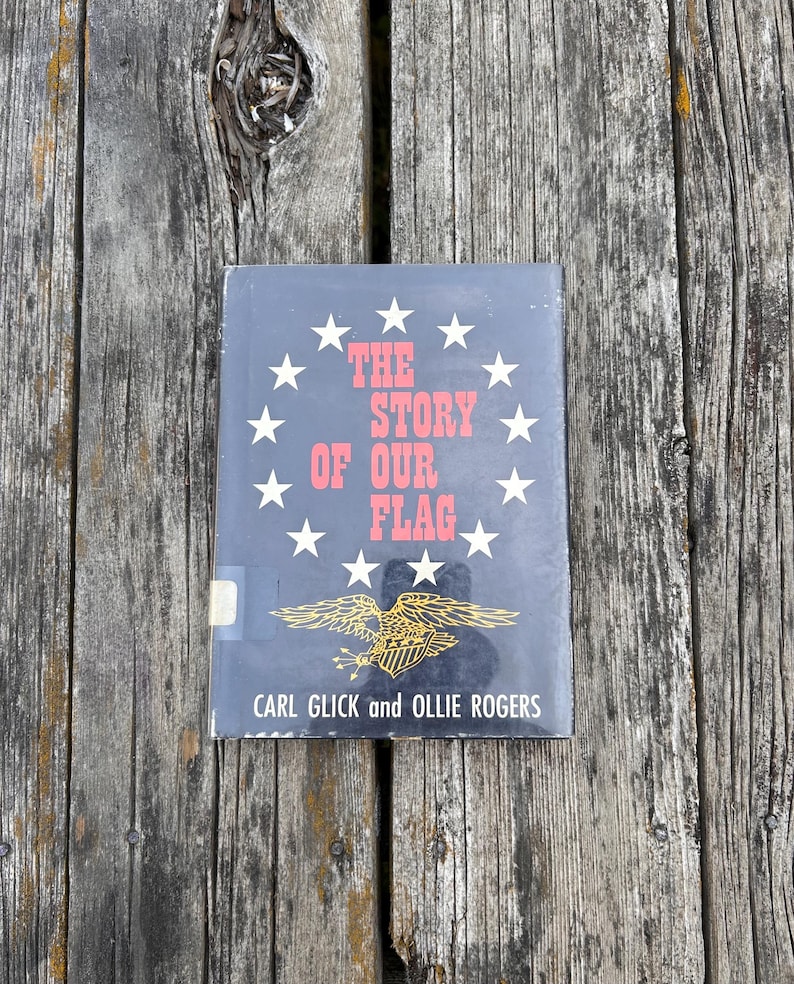 May include: A hardcover book titled The Story of Our Flag by Carl Glick and Ollie Rogers. The black cover features a circle of twelve white stars surrounding the red text, with a gold eagle illustration and a small white library sticker.