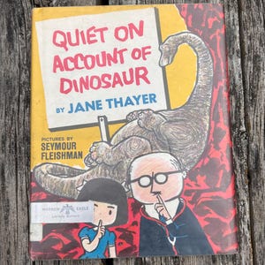 Pode incluir: Um livro de capa dura intitulado Quiet on Account of Dinosaur de Jane Thayer com ilustra&ccedil;&otilde;es de Seymour Fleishman. A capa mostra um grande dinossauro, uma crian&ccedil;a e um adulto de &oacute;culos, todos fazendo um gesto de sil&ecirc;ncio sobre um fundo amarelo.