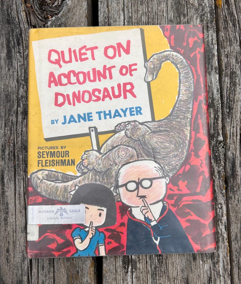 Pode incluir: Um livro de capa dura intitulado Quiet on Account of Dinosaur de Jane Thayer com ilustra&ccedil;&otilde;es de Seymour Fleishman. A capa mostra um grande dinossauro, uma crian&ccedil;a e um adulto de &oacute;culos, todos fazendo um gesto de sil&ecirc;ncio sobre um fundo amarelo.