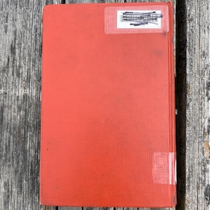May include: A rectangular hardcover book with a solid orange-red fabric cover. The front cover features a white rectangular library sticker near the top with black text and a barcode, and a small piece of clear adhesive tape near the bottom.
