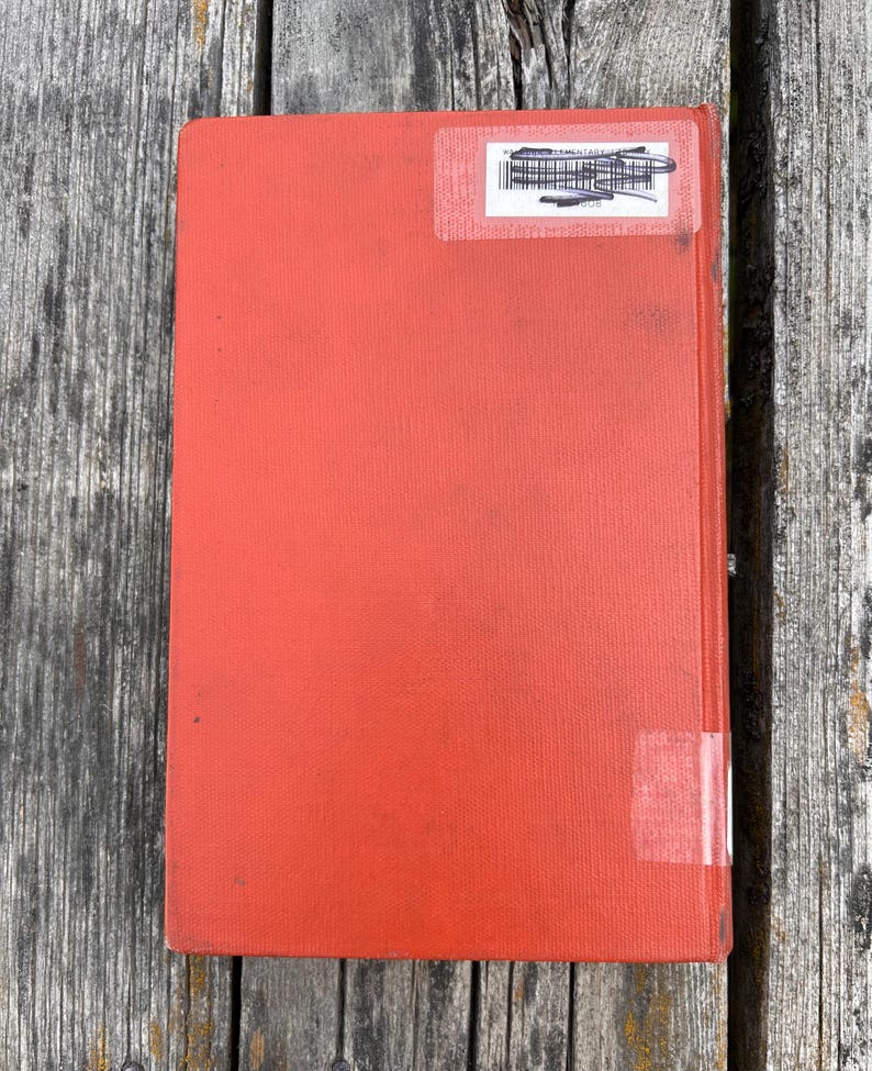 May include: A rectangular hardcover book with a solid orange-red fabric cover. The front cover features a white rectangular library sticker near the top with black text and a barcode, and a small piece of clear adhesive tape near the bottom.