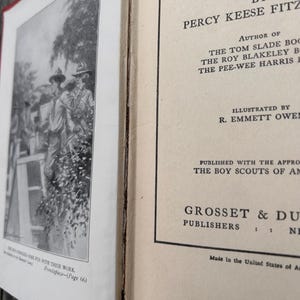 May include: A vintage hardcover book titled Roy Blakeley in the Haunted Camp by Percy Keese Fitzhugh. The open book displays a black and white frontispiece illustration of scouts and a title page with text identifying the author and publisher.