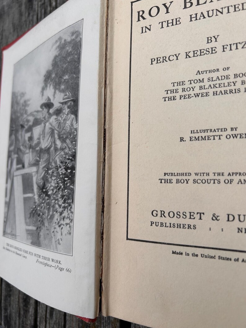May include: A vintage hardcover book titled Roy Blakeley in the Haunted Camp by Percy Keese Fitzhugh. The open book displays a black and white frontispiece illustration of scouts and a title page with text identifying the author and publisher.