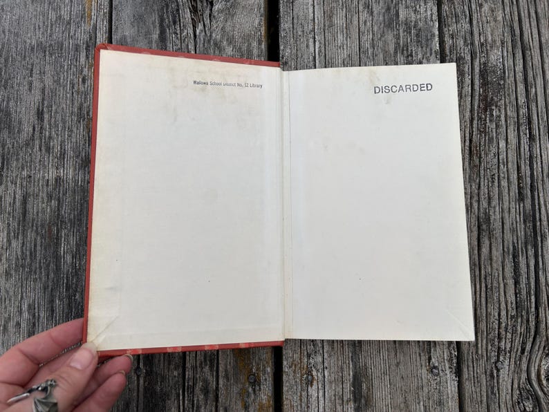 May include: An open hardcover book with a red spine and cover edges. The inside front cover and endpaper are off-white. The text "Wallowa School District No. 12 Library" is printed at the top left, and "DISCARDED" is printed on the right.