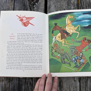 Może przedstawiać: Otwarta książka z tekstem Sir Lancelot's Journey na lewej stronie z ilustracją czerwonej tarczy oraz kolorowym obrazem na prawej stronie przedstawiającym rycerza na białym koniu walczącego z trzema postaciami w zielonym krajobrazie.
