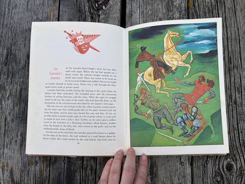 Może przedstawiać: Otwarta książka z tekstem Sir Lancelot's Journey na lewej stronie z ilustracją czerwonej tarczy oraz kolorowym obrazem na prawej stronie przedstawiającym rycerza na białym koniu walczącego z trzema postaciami w zielonym krajobrazie.