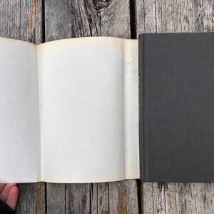 May include: A hardcover book with a solid black cover and a plain, off-white paper dust jacket flap extended to the left. The book is positioned vertically, showing the front cover and the interior side of the dust jacket flap against a wood surface.