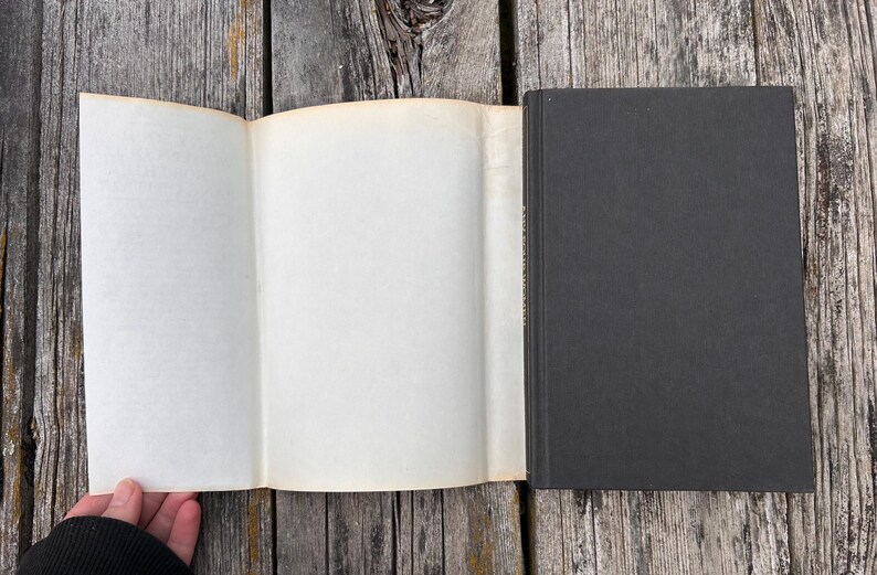 May include: A hardcover book with a solid black cover and a plain, off-white paper dust jacket flap extended to the left. The book is positioned vertically, showing the front cover and the interior side of the dust jacket flap against a wood surface.