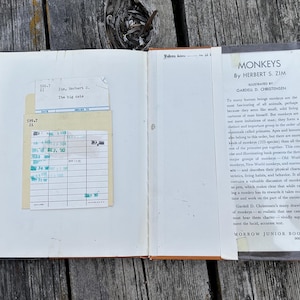 Puede incluir: Un libro de tapa dura abierto titulado Monkeys de Herbert S. Zim, con una sobrecubierta blanca con texto negro y un bolsillo para tarjeta de biblioteca pegado en la cubierta interior con una tarjeta de papel que contiene anotaciones en tinta azul.