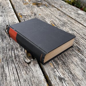 Pu&ograve; includere: Un libro con copertina rigida nera e un accento rosso sul dorso. Il libro presenta pagine di colore chiaro visibili lungo il bordo. Il dorso mostra un piccolo testo di colore dorato stampato verticalmente vicino alla parte inferiore.