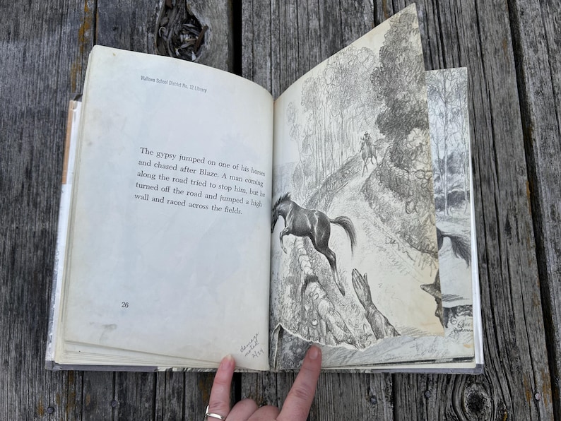 May include: An open vintage book with page 26 visible, featuring black text and a detailed black and white illustration of a horse jumping over a stone wall. The text reads: The gypsy jumped on one of his horses and chased after Blaze.