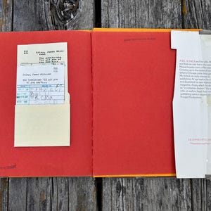 May include: An open hardcover book with bright red endpapers. On the left, a cream-colored library checkout card is tucked into a pocket. On the right, a white dust jacket flap with printed biographical text is attached to the edge of the book cover.