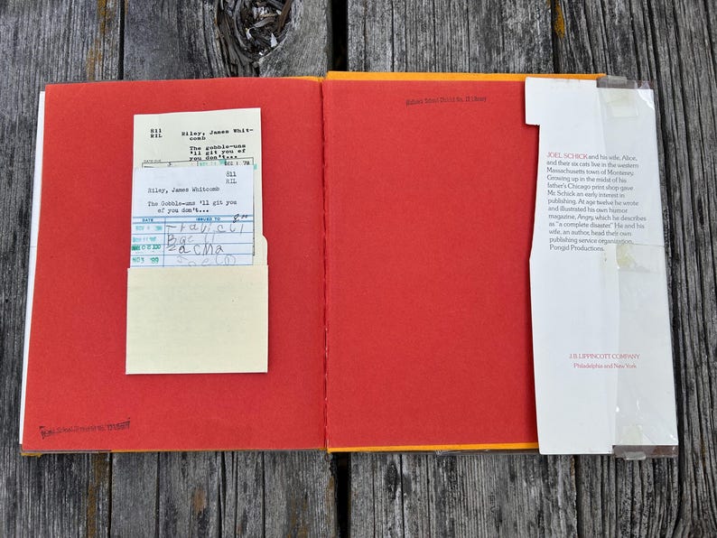 May include: An open hardcover book with bright red endpapers. On the left, a cream-colored library checkout card is tucked into a pocket. On the right, a white dust jacket flap with printed biographical text is attached to the edge of the book cover.