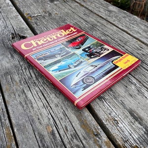 Op de afbeelding: Een rood hardcover boek getiteld Great Cars From Chevrolet by the Editors of Consumer Guide. De kaft bevat vier kleurenfoto's van vintage Chevrolet-auto's en een gele ronde sticker met de tekst Bargain Books Only $.