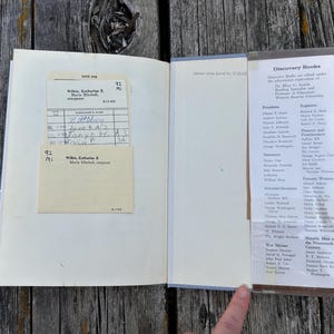 Puede incluir: Un libro de tapa dura abierto con una ficha de pr&eacute;stamo de biblioteca de color crema en un bolsillo en la p&aacute;gina izquierda. La p&aacute;gina derecha presenta una lista impresa titulada Discovery Books, categorizada por figuras hist&oacute;ricas.