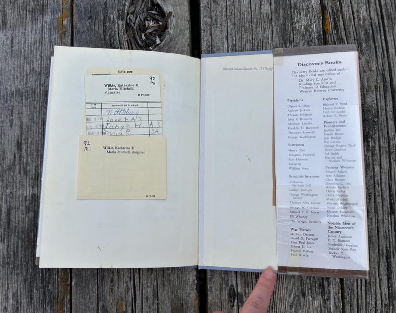 Puede incluir: Un libro de tapa dura abierto con una ficha de pr&eacute;stamo de biblioteca de color crema en un bolsillo en la p&aacute;gina izquierda. La p&aacute;gina derecha presenta una lista impresa titulada Discovery Books, categorizada por figuras hist&oacute;ricas.