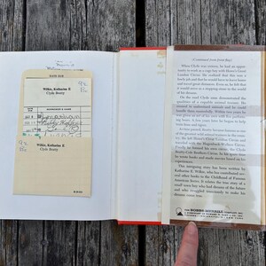 May include: An open hardcover book with a cream-coloured library checkout card in a pocket on the left page. The right page features a printed summary of the biography of Clyde Beatty, published by The Bobbs-Merrill Company, Inc.
