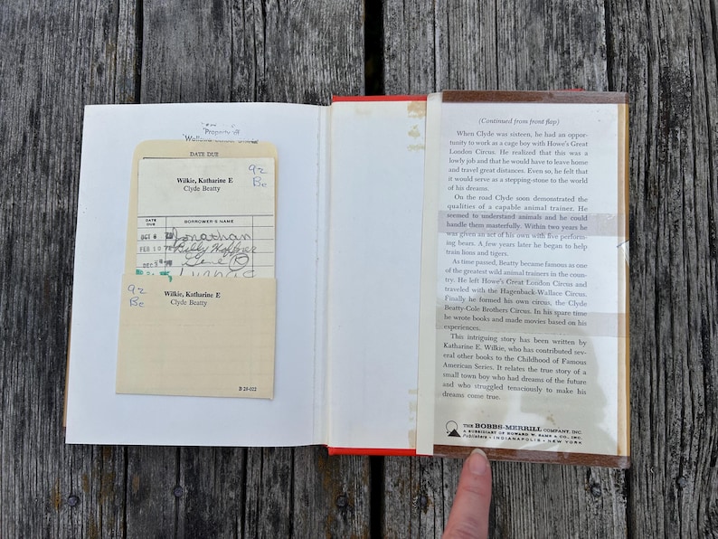 May include: An open hardcover book with a cream-coloured library checkout card in a pocket on the left page. The right page features a printed summary of the biography of Clyde Beatty, published by The Bobbs-Merrill Company, Inc.