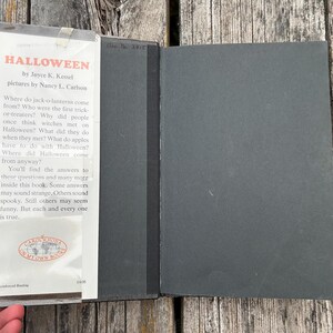 Może przedstawiać: Książka w twardej oprawie zatytułowana Halloween autorstwa Joyce K. Kessel z ilustracjami Nancy L. Carlson. Książka posiada białą klapkę obwoluty z czarnym tekstem i czerwonym tytułem, przymocowaną do gładkiej czarnej okładki. Tekst opisuje historię.