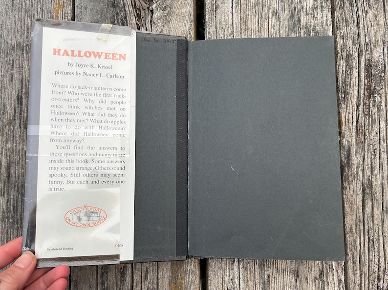 Może przedstawiać: Książka w twardej oprawie zatytułowana Halloween autorstwa Joyce K. Kessel z ilustracjami Nancy L. Carlson. Książka posiada białą klapkę obwoluty z czarnym tekstem i czerwonym tytułem, przymocowaną do gładkiej czarnej okładki. Tekst opisuje historię.