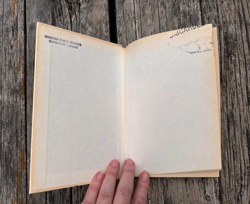 Pu&ograve; includere: Un libro con copertina rigida aperto con pagine color crema. La pagina sinistra presenta un timbro a inchiostro nero con la scritta WALLOWA PUBLIC SCHOOL ELEMENTARY LIBRARY. La pagina destra ha un timbro nero nell'angolo in alto a destra con la scritta DISCARDED.