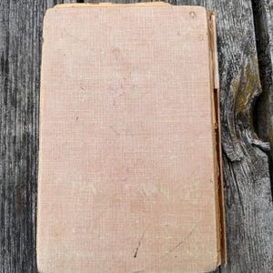 K&ouml;nnte beinhalten: Ein rechteckiges Hardcover-Buch mit einem verblassten, hellbeigen, strukturierten Stoffeinband. Die Kanten des Einbands weisen Abnutzungserscheinungen, Ausfransungen und Verf&auml;rbungen auf, mit sichtbaren Kratzern und kleinen dunklen Flecken.