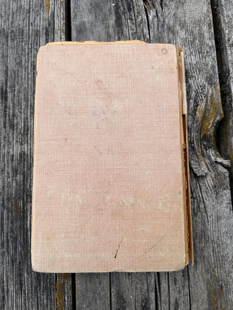 K&ouml;nnte beinhalten: Ein rechteckiges Hardcover-Buch mit einem verblassten, hellbeigen, strukturierten Stoffeinband. Die Kanten des Einbands weisen Abnutzungserscheinungen, Ausfransungen und Verf&auml;rbungen auf, mit sichtbaren Kratzern und kleinen dunklen Flecken.