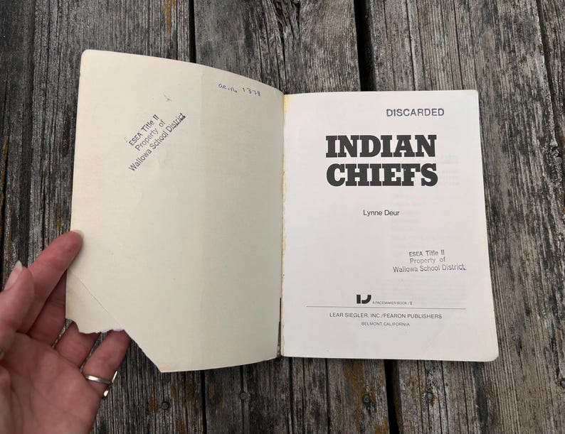 Może przedstawiać: Otwarta książka pod tytułem Indian Chiefs autorstwa Lynne Deur, wydana przez Lear Siegler, Inc./Fearon Publishers. Na wewnętrznej stronie okładki znajduje się pieczątka ESEA Title II Property of Wallowa School District oraz nadrukowane słowo DISCARDED.
