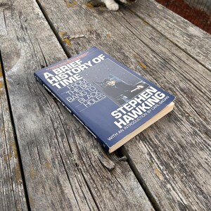 Peut inclure: Un livre de Stephen Hawking, intitul&eacute; "Une br&egrave;ve histoire du temps", avec une introduction de Carl Sagan. La couverture est bleu fonc&eacute; avec du texte blanc et une photo. Le livre est pos&eacute; sur une surface en bois vieilli.