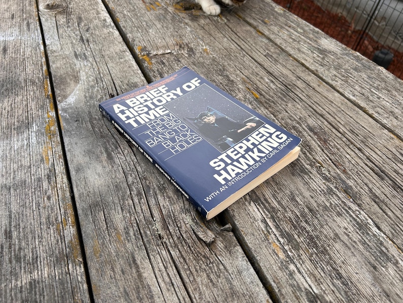 Peut inclure: Un livre de Stephen Hawking, intitul&eacute; "Une br&egrave;ve histoire du temps", avec une introduction de Carl Sagan. La couverture est bleu fonc&eacute; avec du texte blanc et une photo. Le livre est pos&eacute; sur une surface en bois vieilli.