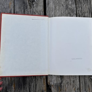 May include: An open hardcover book with a red spine and white endpapers. The left page features small black text reading 'Mullins School District No. 15 Library' at the top. The right page has the word 'DISCARDED' stamped in black ink.